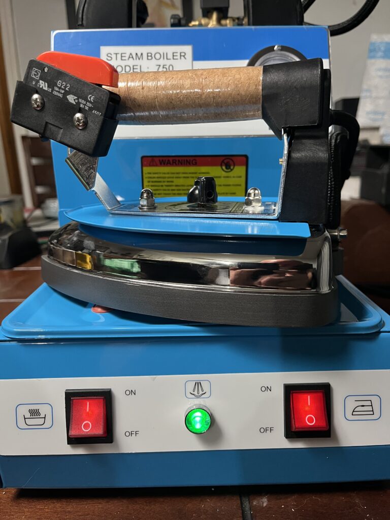 ST-750 heated steam iron resting on boiler unit showing handle, temperature control, steam control, and connections